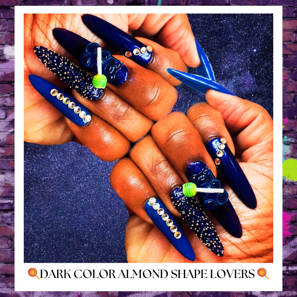 Close-up of deep navy blue stiletto press-on nails with rhinestone accents, textured glitter detailing, and gummy candy lollipop charms on a sparkling dark background.