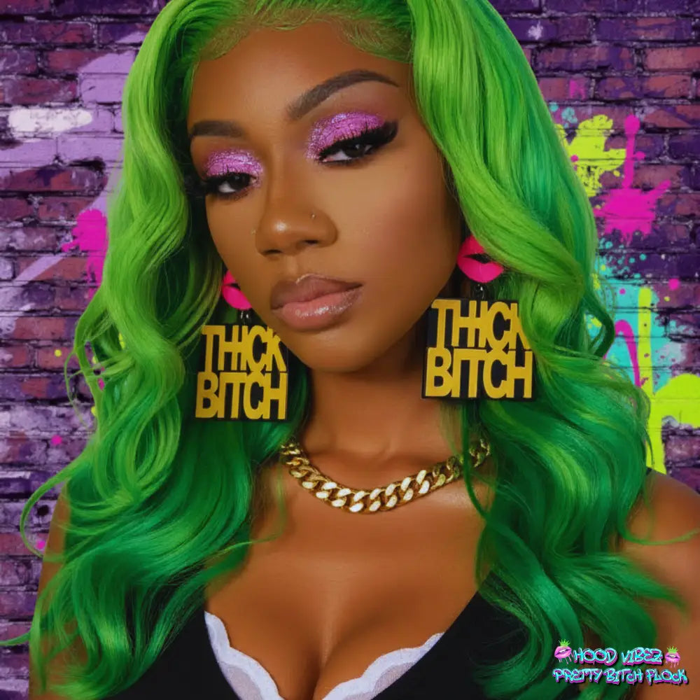 Thick Bitch statement earrings featuring bold gold lettering on black acrylic with pink lip accents against a glittering gold and green background.