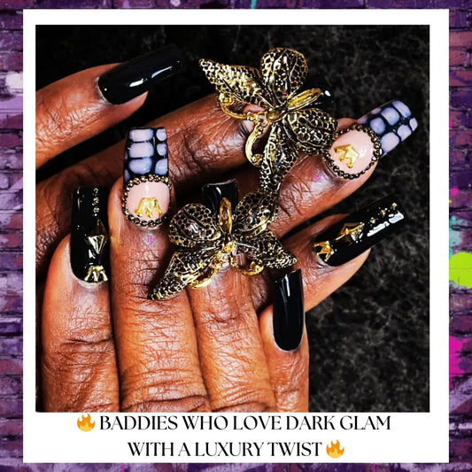 Close-up of luxury black press-on nails with glossy black polish, gold butterfly charms, rhinestone accents, and marble-style details on hands against a dark textured background.