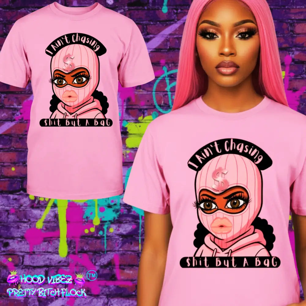 Model wearing “Pink t-shirt with cartoon baddie wearing a pink ski mask and hoodie, with text reading ‘I Ain’t Chasing Shit But a Bag’ in black and pink.”