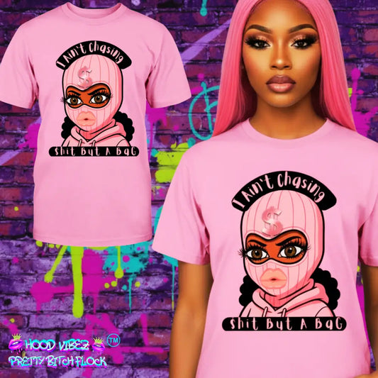 Model wearing “Pink t-shirt with cartoon baddie wearing a pink ski mask and hoodie, with text reading ‘I Ain’t Chasing Shit But a Bag’ in black and pink.”