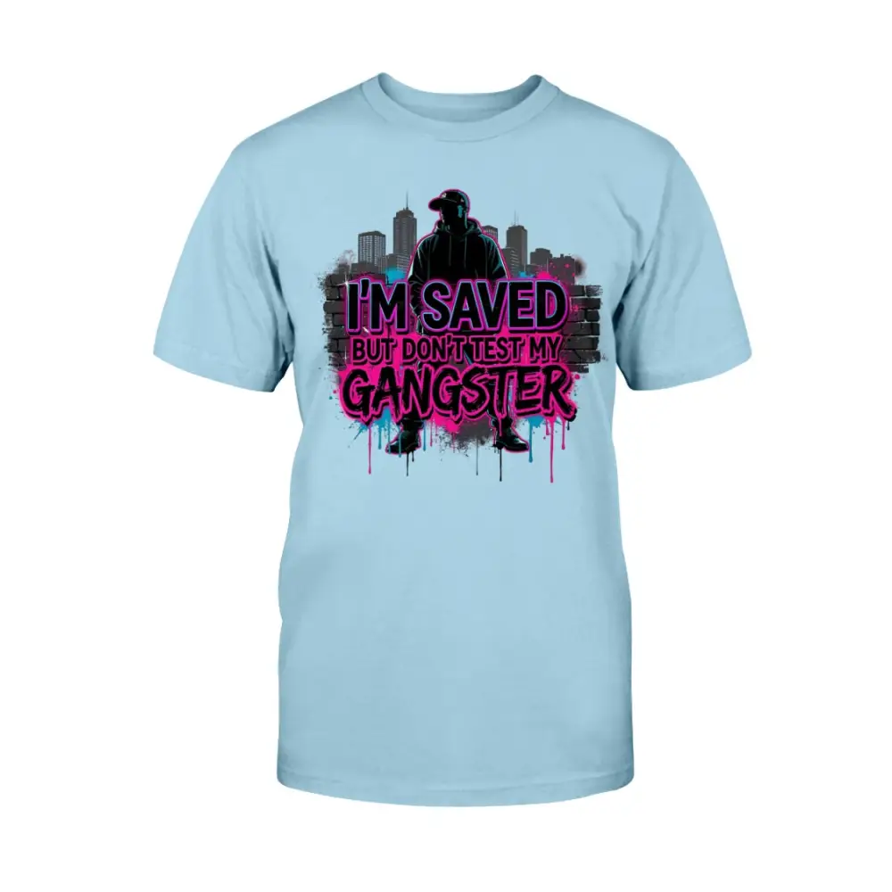 I’m Saved But Don’t Test My Gangster graphic t-shirt featuring bold pink graffiti lettering, urban skyline background, and streetwear silhouette design on light blue tee.