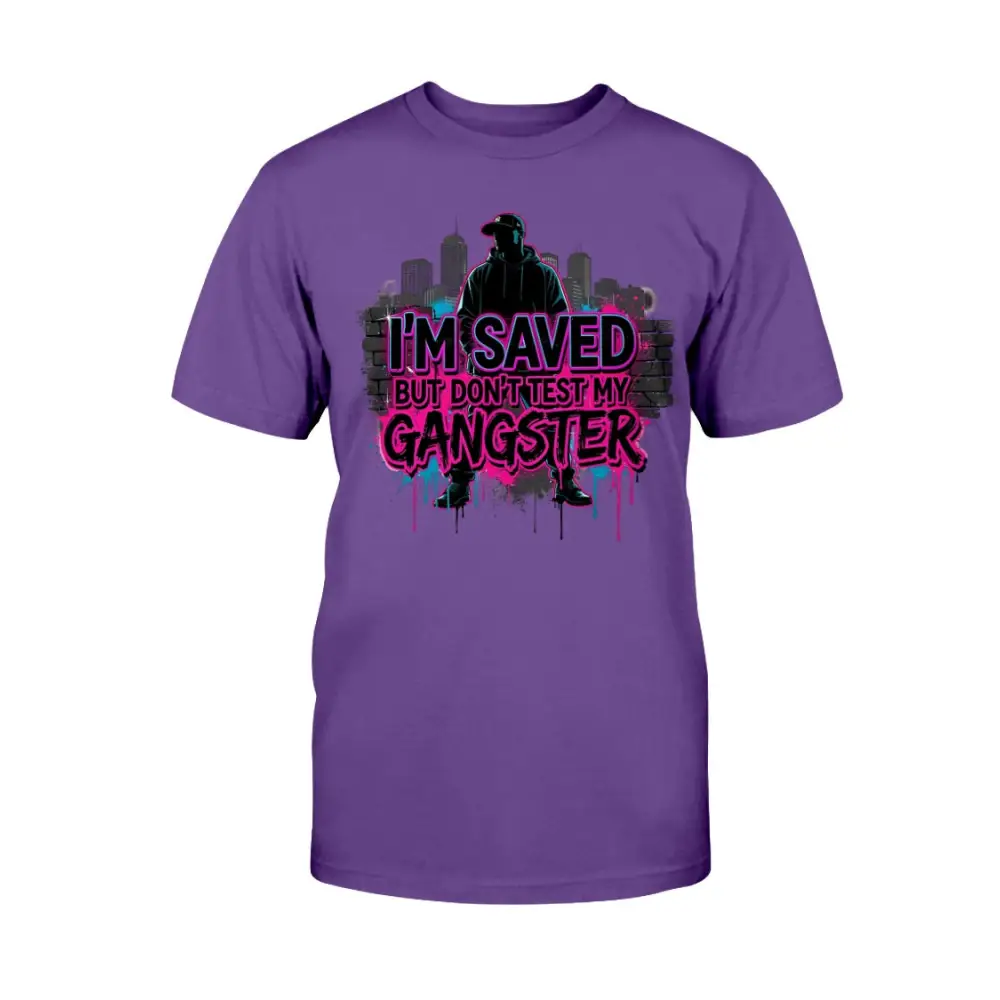 I’m Saved But Don’t Test My Gangster graphic t-shirt featuring bold pink graffiti lettering, urban skyline background, and streetwear silhouette design on purple tee.