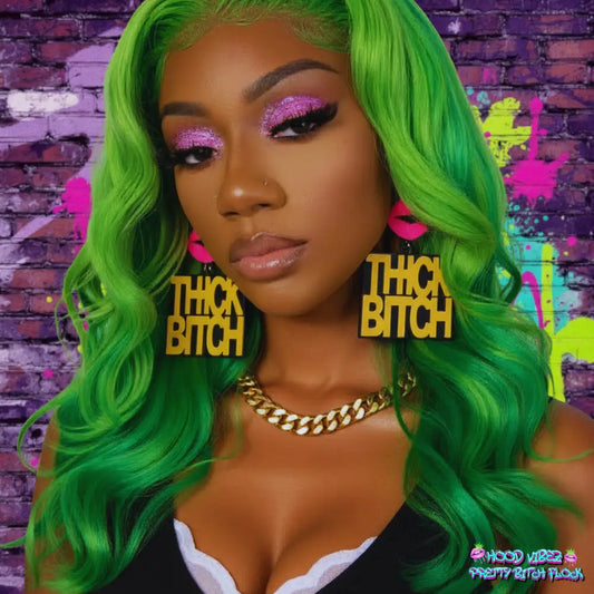 Thick Bitch statement earrings featuring bold gold lettering on black acrylic with pink lip accents against a glittering gold and green background.